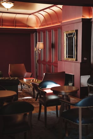 A dimly lit room with a curved ceiling, a red wall, and a wooden floor. There are several chairs and a table with a lamp.