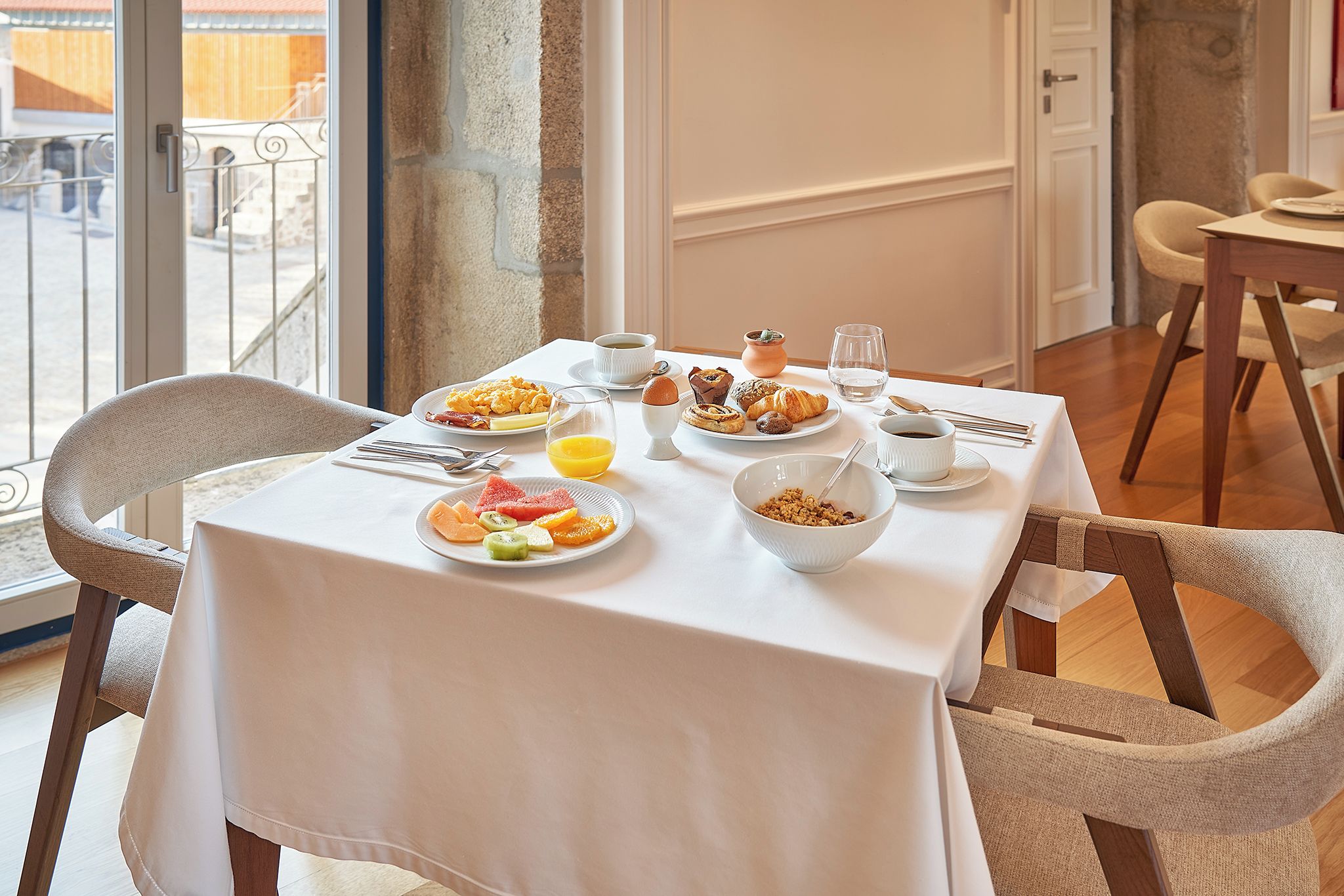 A dining table set with breakfast items, including fruits, pastries, and beverages, in a room with a view of the outdoors.
