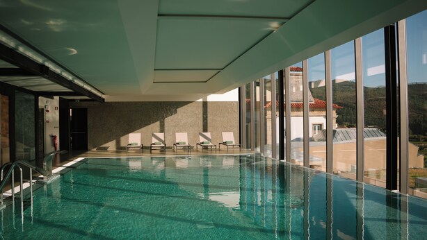 A modern indoor swimming pool with clear water, sunlit by large windows, and a view of a hilly landscape. - The Vinea Collection Hotel (Monção)