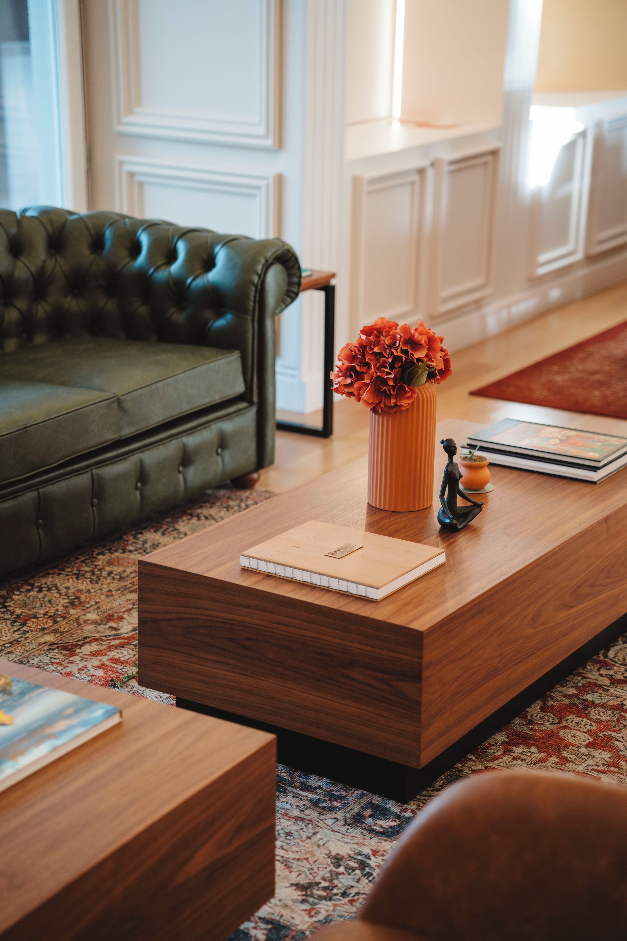 A modern living room with a green sofa, a wooden coffee table with a notebook, a vase with flowers, and a small statue.