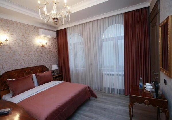 A hotel room with a large bed, a wooden nightstand, a desk with a chair, a chandelier, and a large window with curtains. - West Inn Palace Hotel (Baku)
