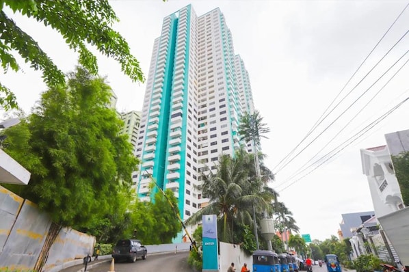 Un edificio alto y moderno con un elemento vertical destacado, rodeado de vegetación e infraestructura urbana.