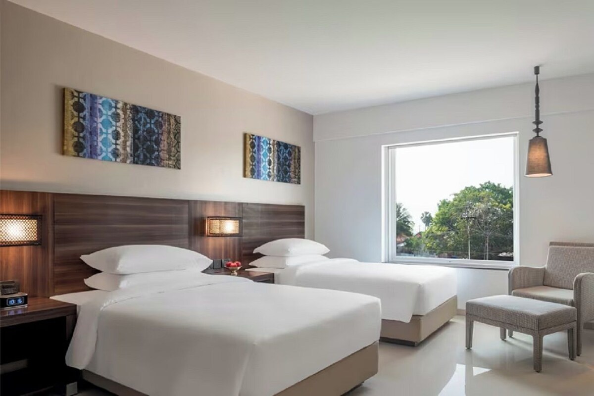 A hotel room with two beds, a television, a chair, artwork on the walls, and a large window.