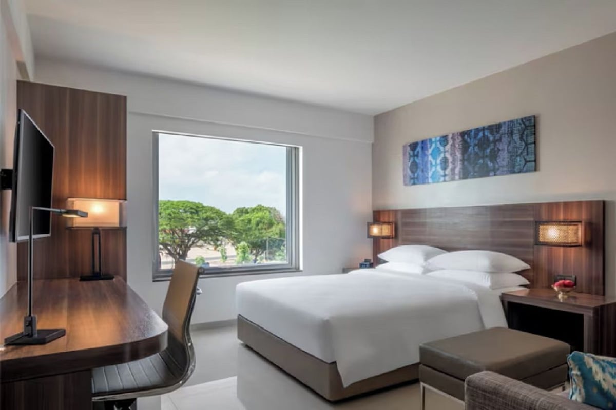 A hotel room with a bed, a desk with a chair, a TV, and a window with a view of trees.