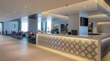 A modern hotel lobby with a reception desk, seating area, and a large window providing natural light.