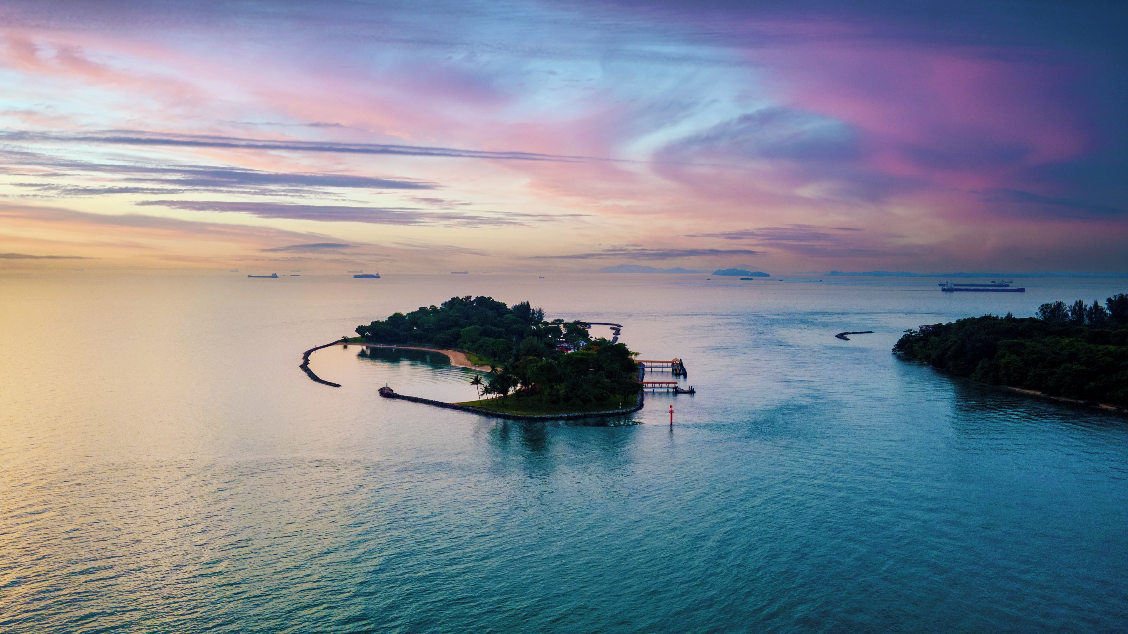An aerial view of a tropical island with a pier, surrounded by calm waters and distant ships.