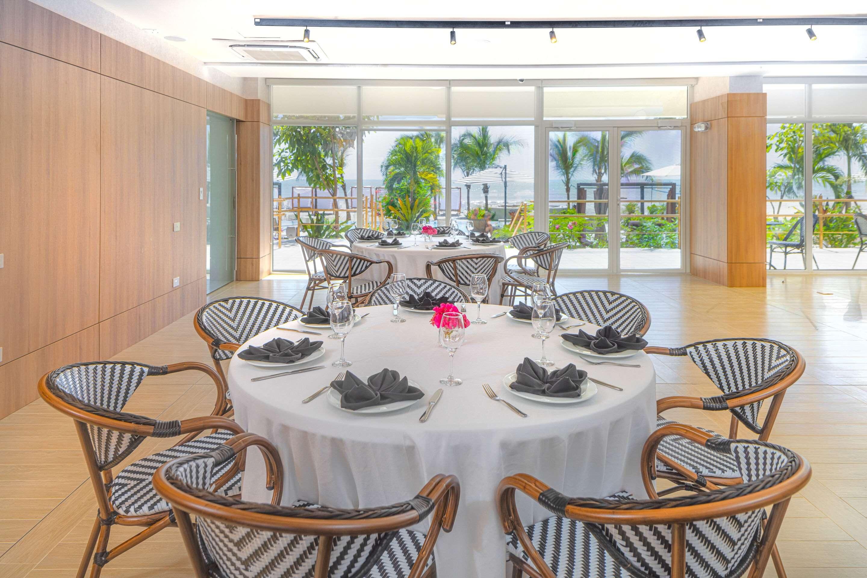 A dining area with round tables, wicker chairs, and a view of the ocean.
