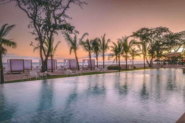 A poolside area with lounge chairs and umbrellas, overlooking a beach at sunset.