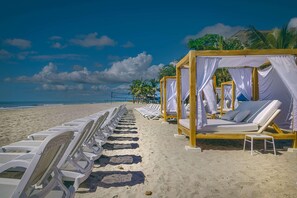 A beach with rows of white sun loungers and wooden cabanas with white curtains.