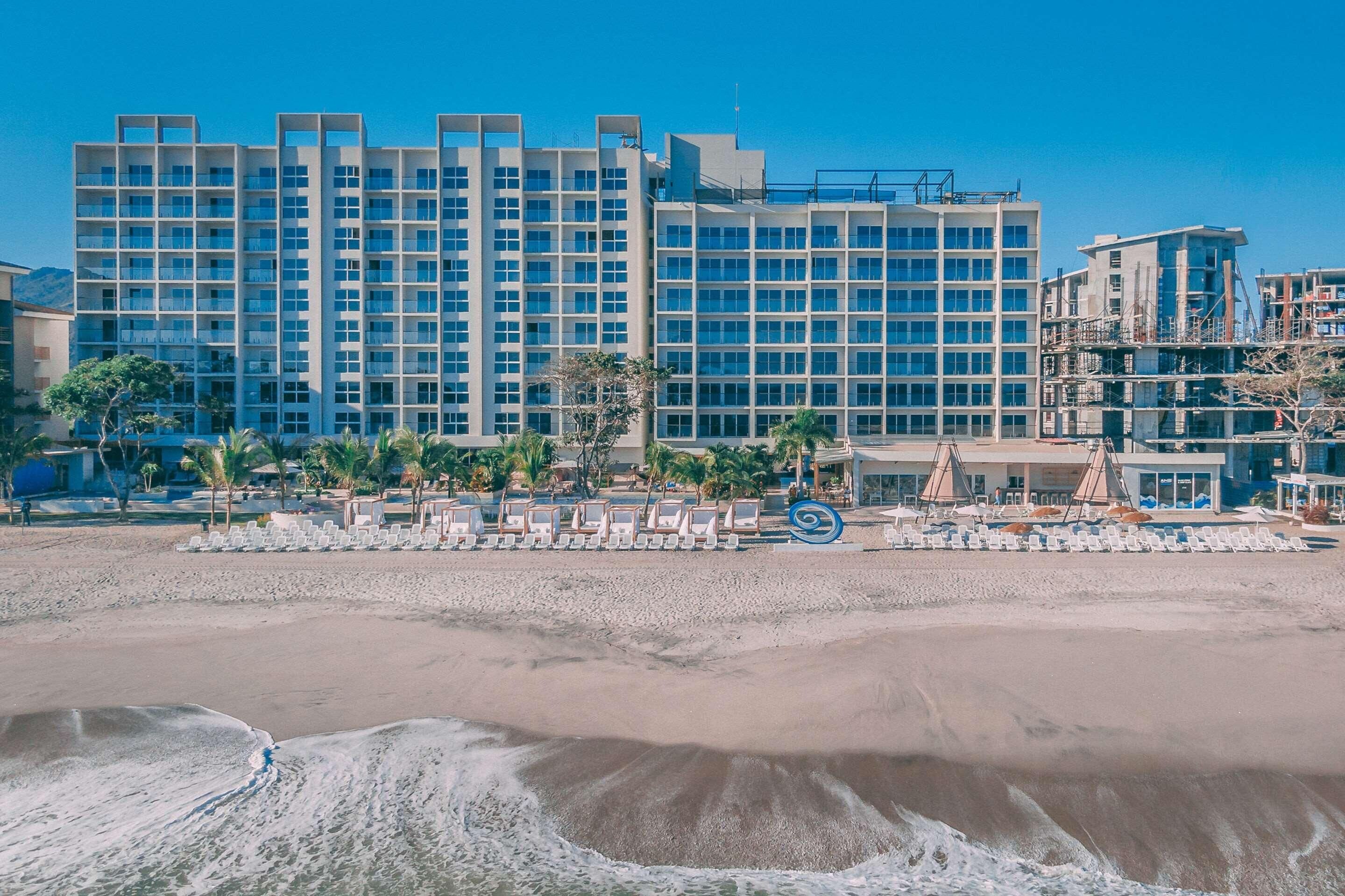 A beachfront hotel with multiple floors, a sandy beach in the foreground, and a clear blue sky.