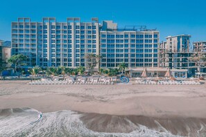A beachfront hotel with multiple floors, a sandy beach in the foreground, and a clear blue sky.