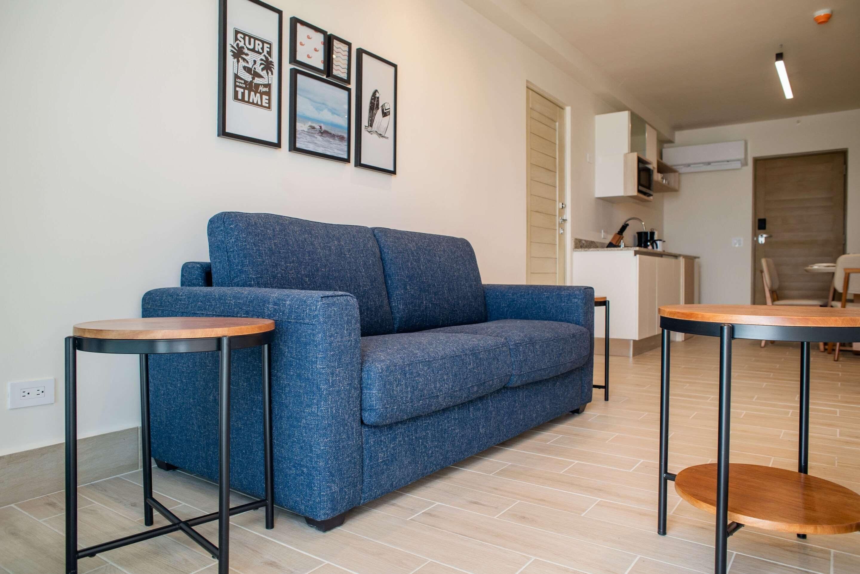 Un salón moderno con un sofá azul, dos mesitas de madera y una cocina al fondo.