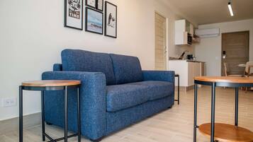 Un salón moderno con un sofá azul, dos mesitas de madera y una cocina al fondo.