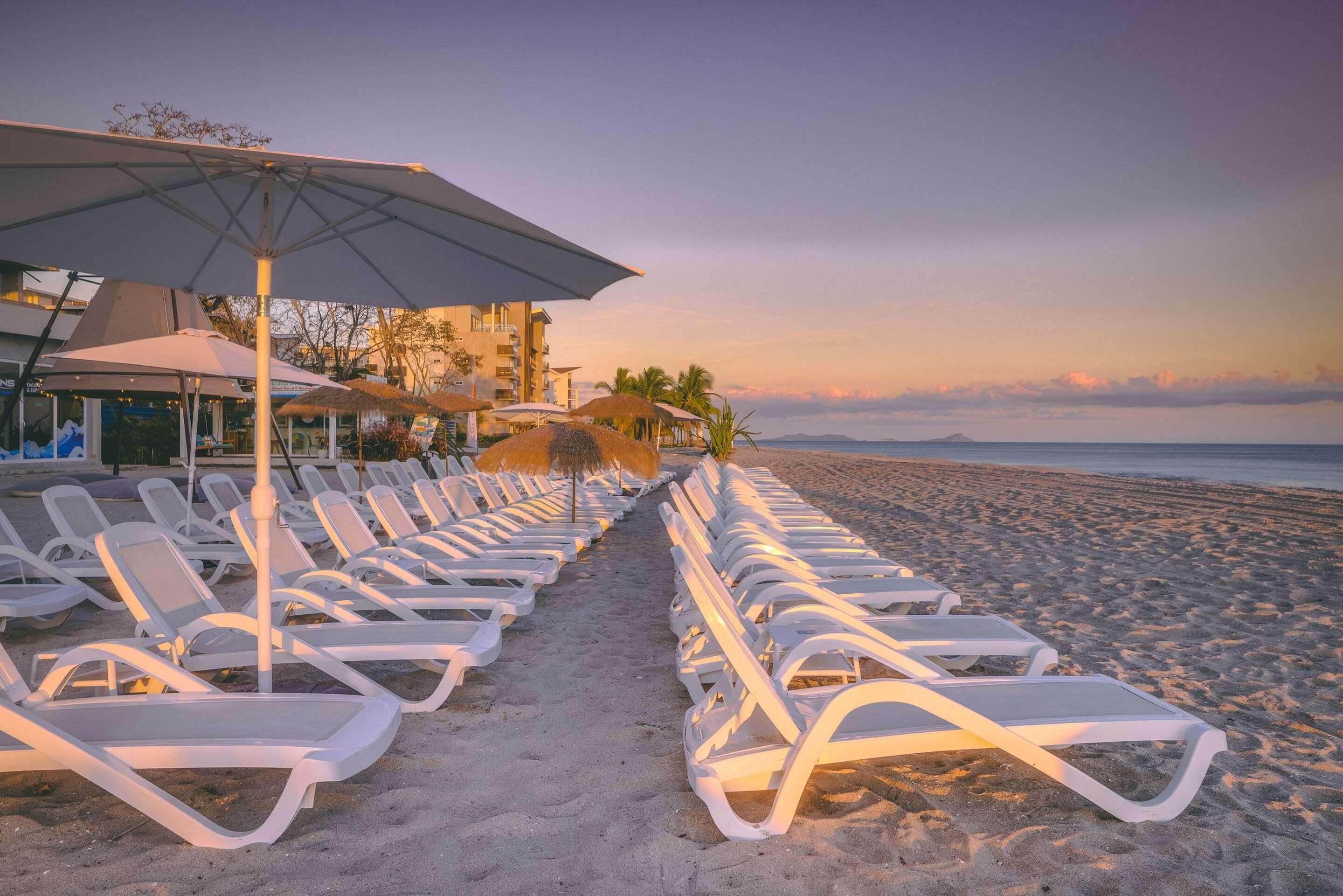 Un hotel frente a la playa con filas de tumbonas y sombrillas, una vista despejada del océano y un edificio con balcón que da a la playa.