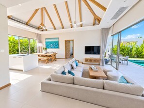 A modern living room with a large sofa, a dining area, and a TV. - Brand New Private 3-bedroom Pool Villa in Bang Tao, Phuket (Thep Krasattri)