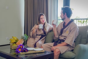 Two people sitting on a couch with a table in front of them holding mugs, a fruit basket, and a tray with plates.