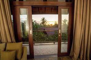 A room with a sliding glass door leading to a balcony with a view of palm trees and a sunset.