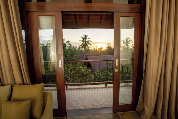 A room with a sliding glass door leading to a balcony with a view of palm trees and a sunset.