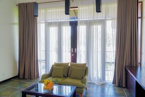 A living room with a sofa, a glass coffee table, and a view through large windows with curtains.