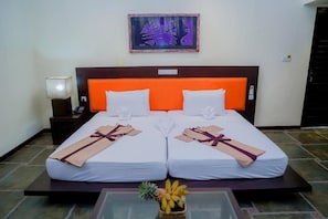 A double bed with white and orange bedding, a bedside lamp, a framed artwork on the wall, and a fruit basket on the floor.