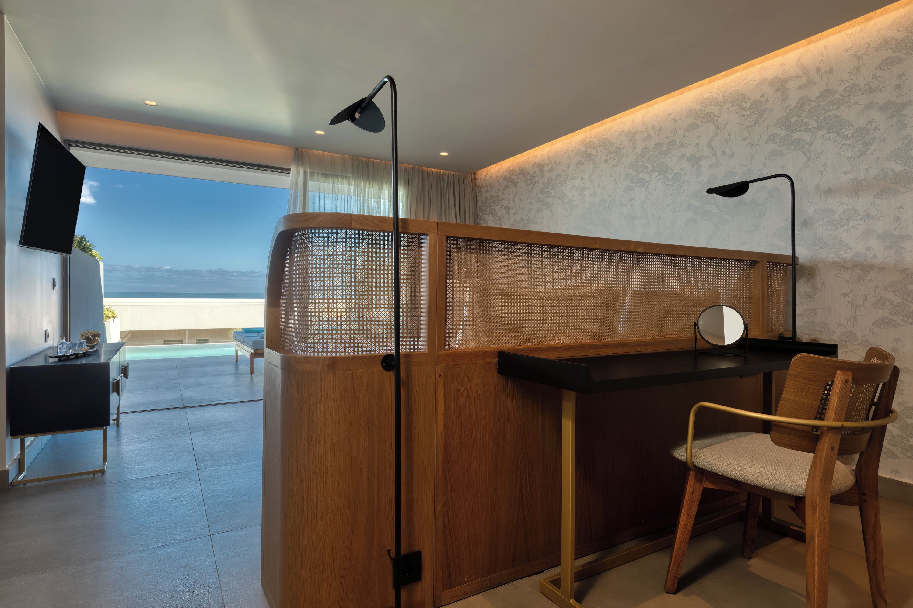 A modern hotel room with a wooden reception desk, a chair, a TV, and a large window offering a view of the ocean.