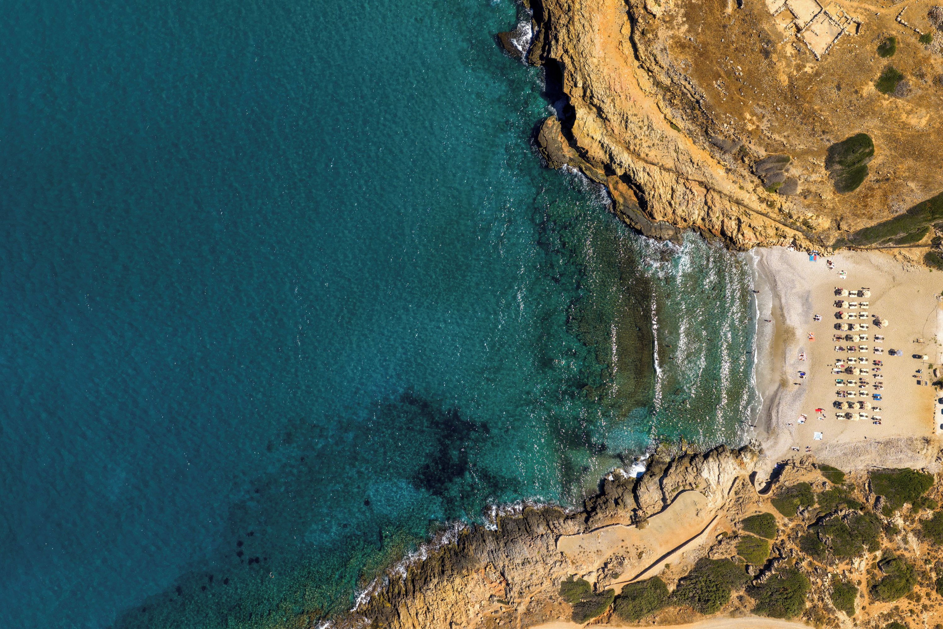 An aerial view of a beach with clear turquoise waters, rocky coastline, and a sandy area with rows of beach chairs and umbrellas.