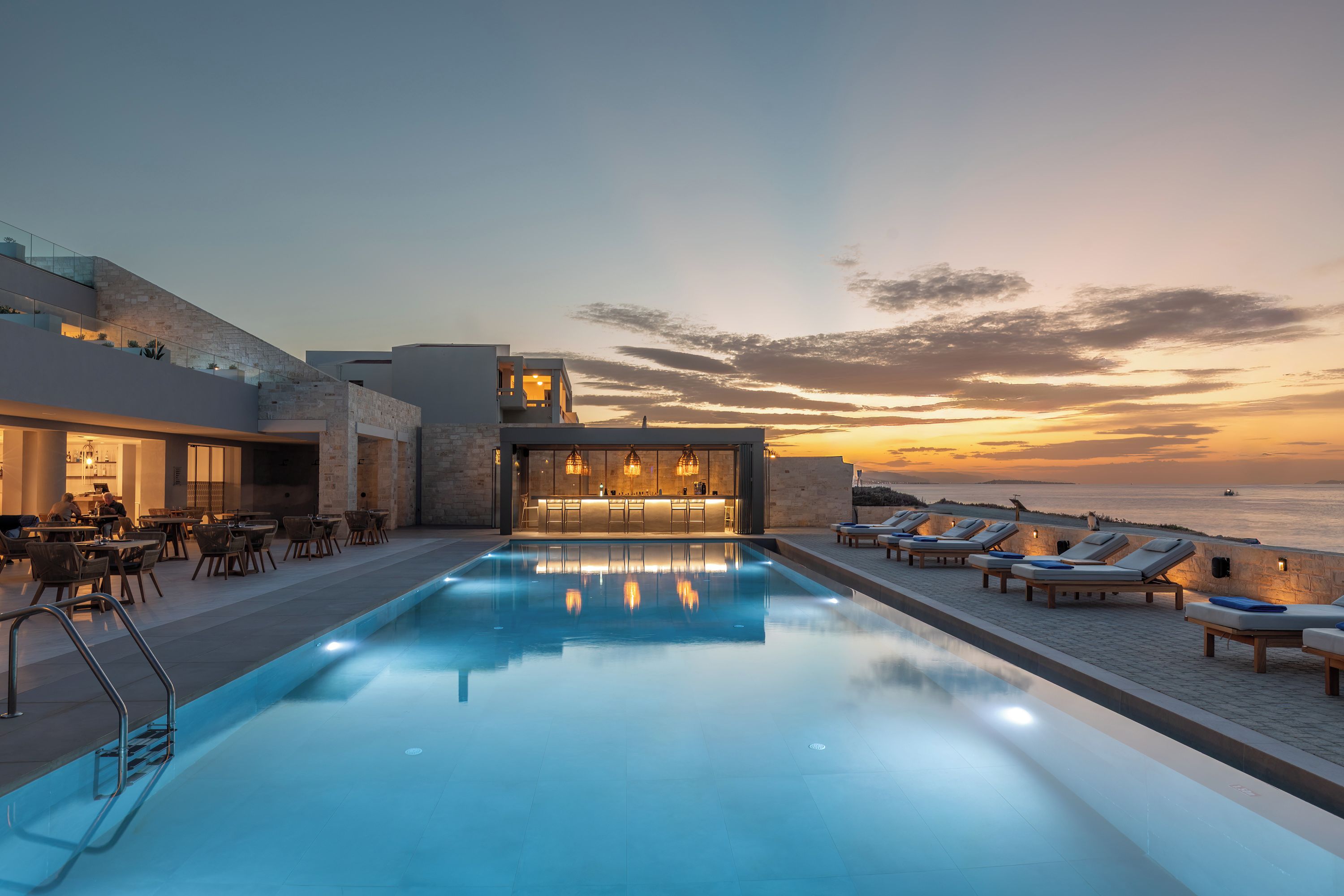 Zona de piscina con sillones de descanso y un bar, con vista a una extensión de agua al atardecer.