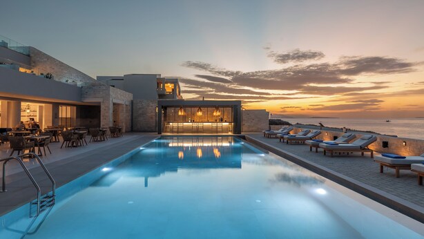Un espace aménagé au bord de la piscine, avec des chaises longues et un bar, offrant une vue sur une étendue d’eau au coucher du soleil.