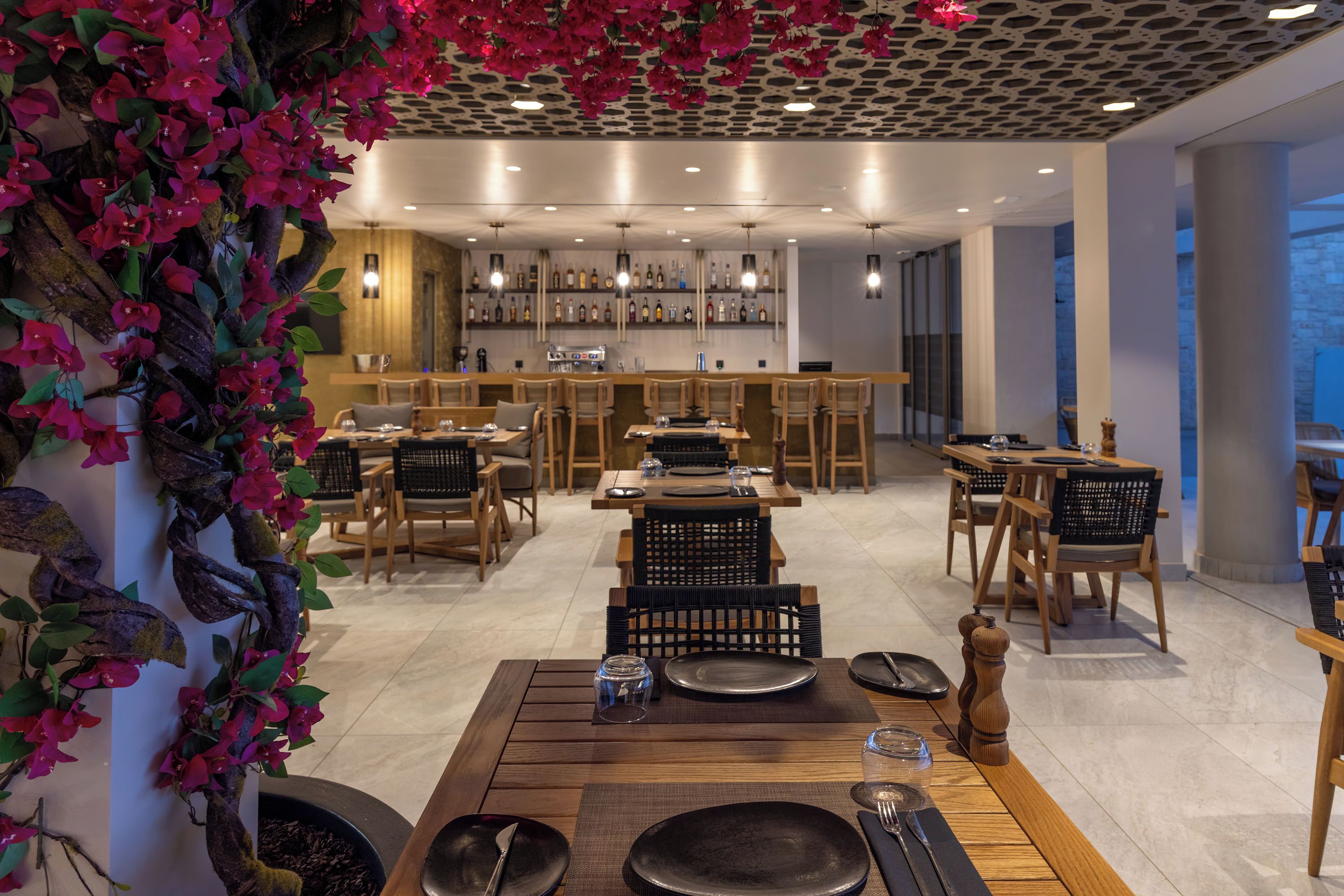 A restaurant with wooden tables and chairs, a bar area with shelves of bottles, and a decorative plant with pink flowers.