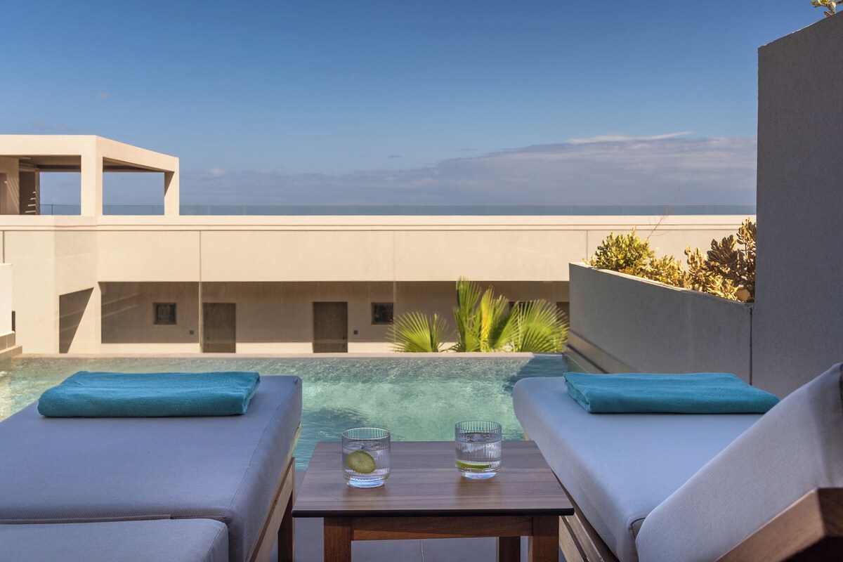 Zona de piscina con dos sillas de descanso y una mesa, con vistas a una piscina cristalina y una vista distante del océano.