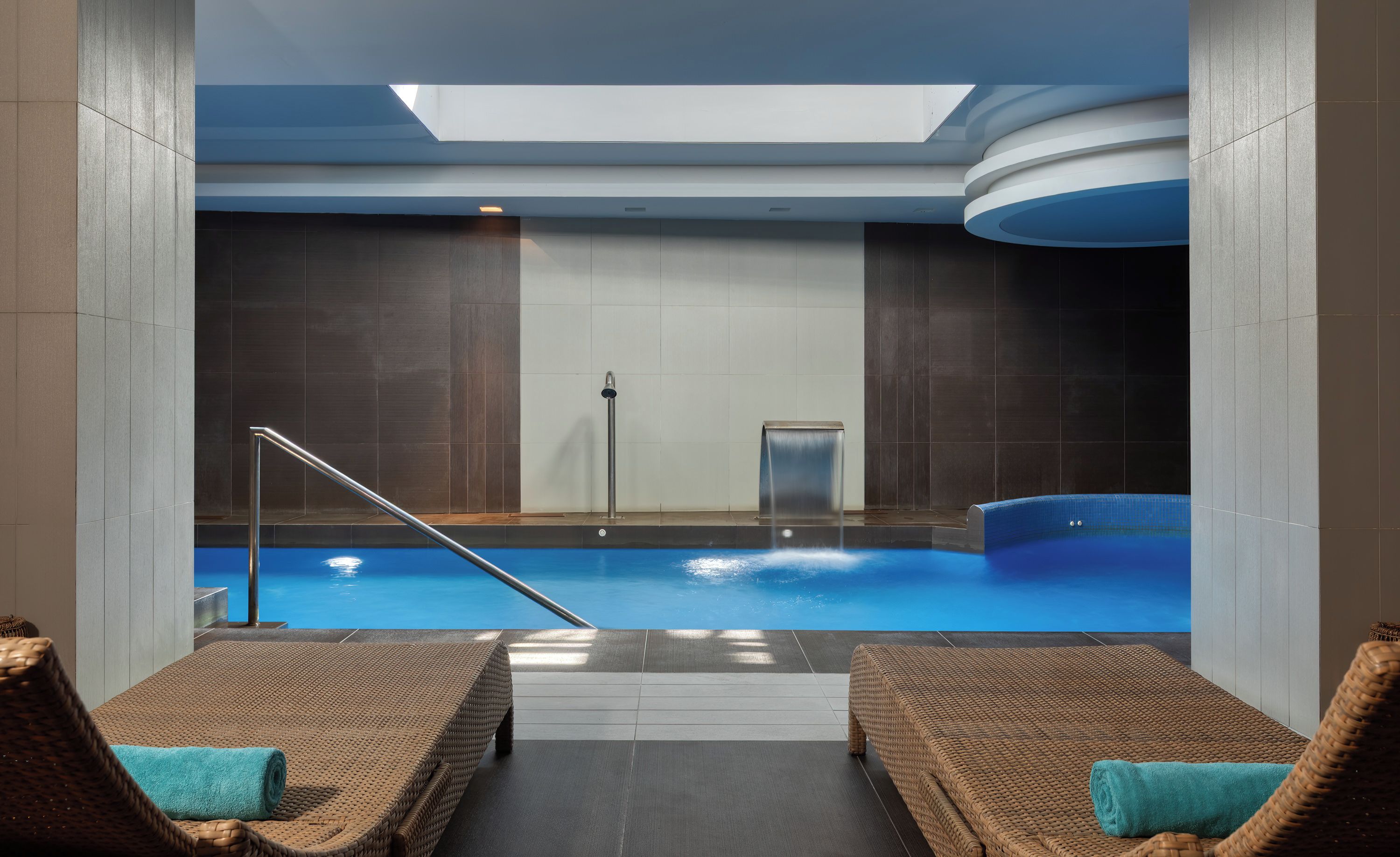 Un área de piscina interior moderna con tragaluces, una fuente de agua y dos sillones de mimbre con toallas.