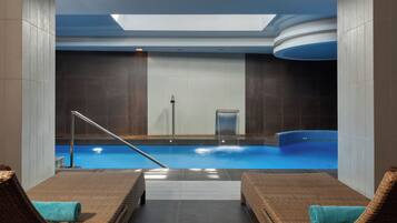A modern indoor pool area with a skylight, a water fountain, and two wicker lounge chairs with towels.
