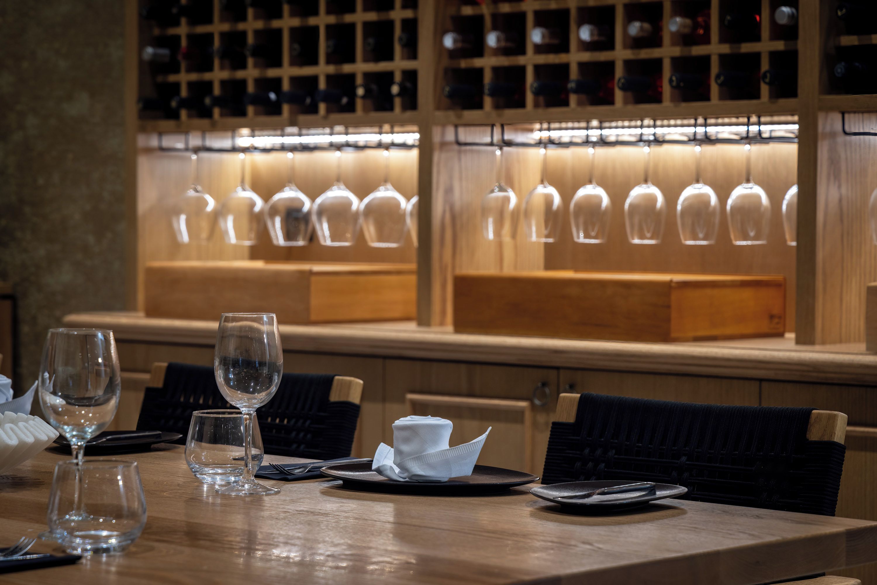 A dining table set with wine glasses, plates, and cutlery, with a wine rack in the background.