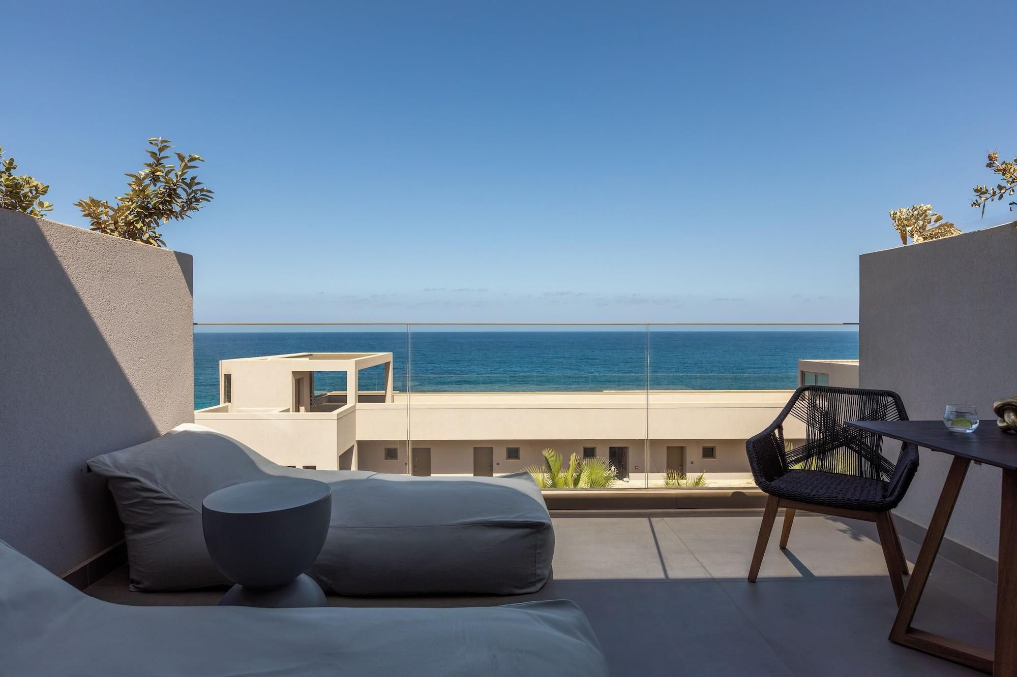 A balcony with a view of the sea, a table with chairs, and a sofa with cushions.
