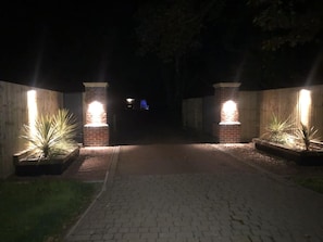 Ein Weg führt zu einem Gebäudeeingang mit beleuchteten Ziegelstützen und Pflanzen in Kübeln.