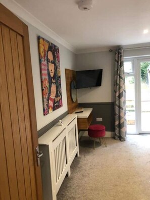 Ein Zimmer mit Holztür, Heizkörper, Schreibtisch, Fernseher und einem roten Hocker.