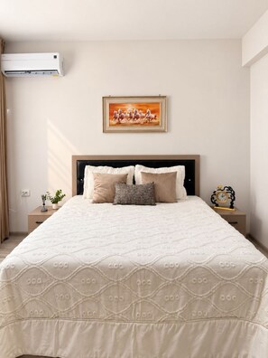 A bedroom with a large bed, a headboard, a bedside table with a clock, a wall-mounted air conditioner, and a framed picture above the bed. - Zarafshan 60 (Tashkent)