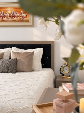 A neatly made bed with a white duvet and several pillows, a bedside table with a clock and books, and a vase with flowers. - Zarafshan 60 (Tashkent)