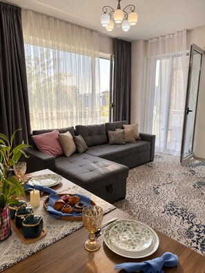 A modern living room with a sectional sofa, a dining table set with plates and a candle, and a large window with sheer curtains. - Zarafshan 60 (Tashkent)