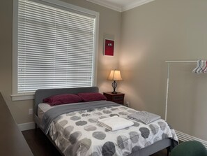 Ein Schlafzimmer mit einem Bett, einem Nachttisch mit Lampe, einem Fenster mit Jalousien, einem Wandhaken für Kleidung und einem gerahmten Bild.