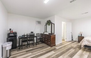 Un dormitorio con cama, cómoda, escritorio con silla, microondas y un cubo de basura.
