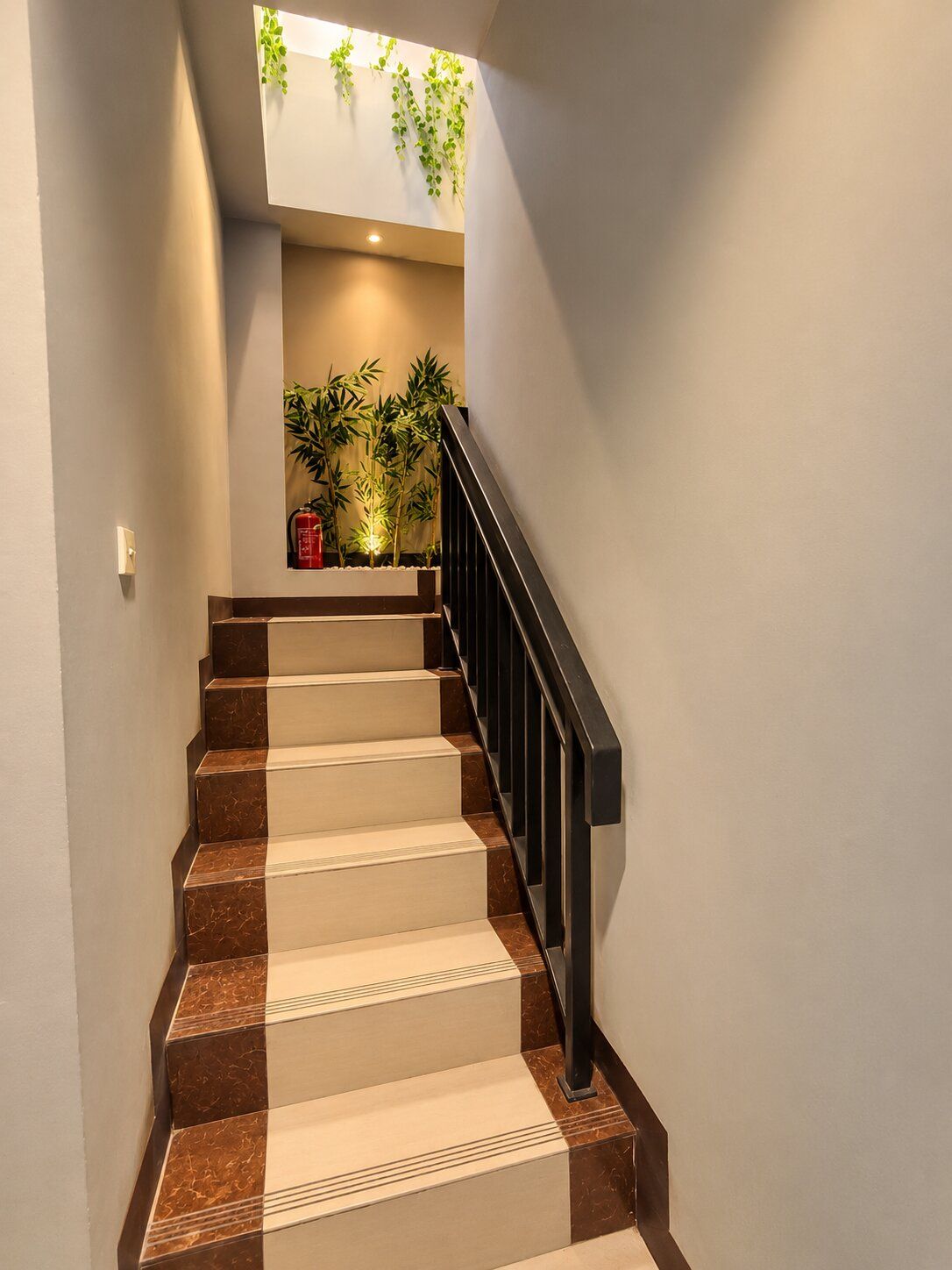 Uma escadaria com degraus alternados em bege e castanho, corrimão preto e uma planta no topo, fixada na parede.