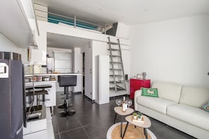 A modern apartment with a kitchen, dining area, and living room. There is a loft space above the kitchen, a red cabinet, and a white sofa with decorative pillows. - Cap Agde Waterfront (Agde)
