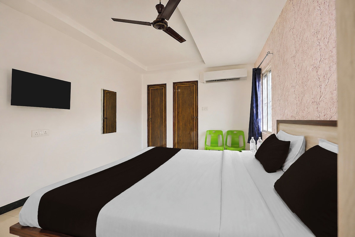 A hotel room with a bed, a ceiling fan, a flat-screen TV, two doors, an air conditioning unit, and two green chairs.