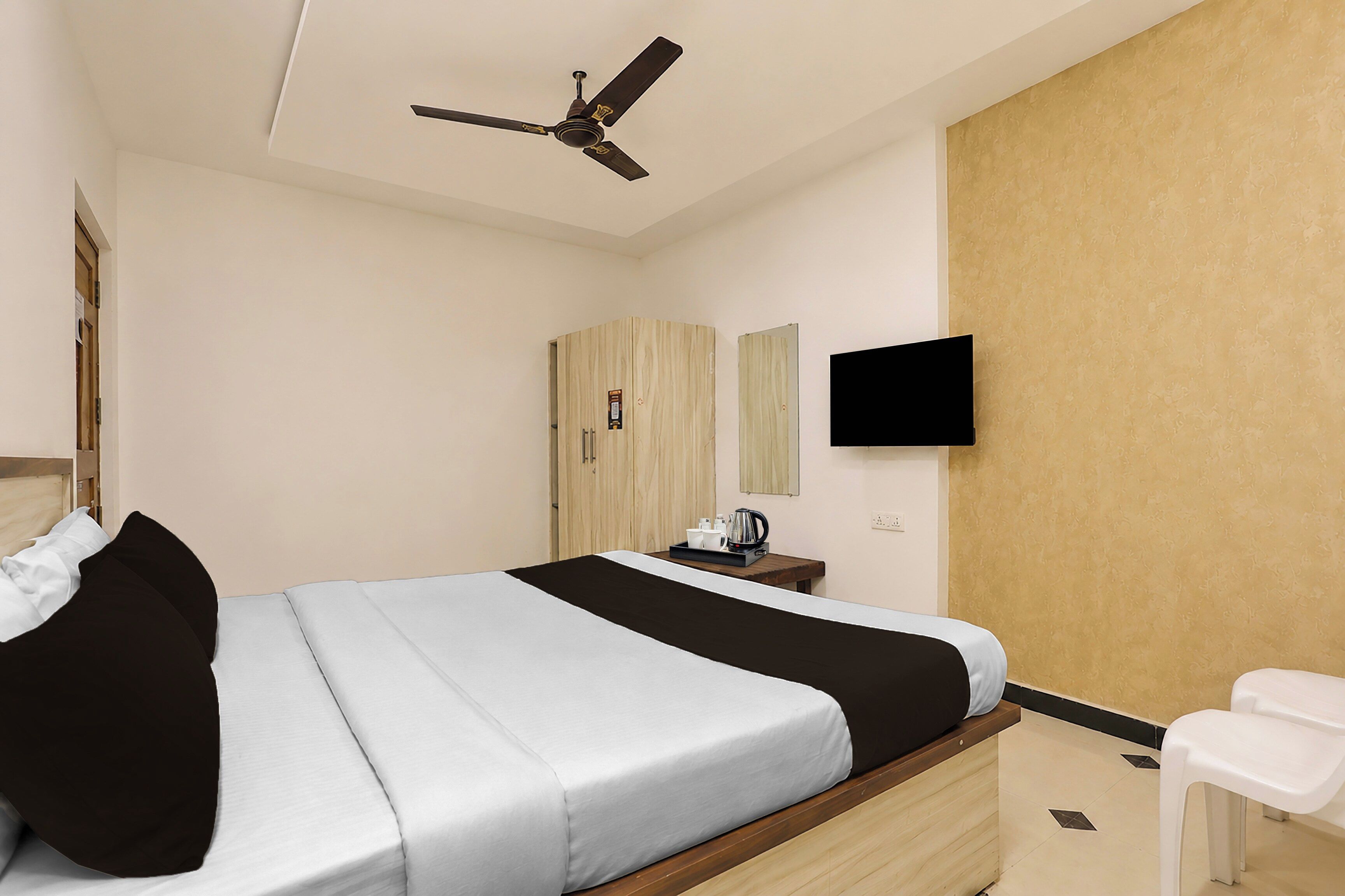 Habitación de hotel con cama, escritorio con cafetera, televisor en la pared y ventilador de techo.