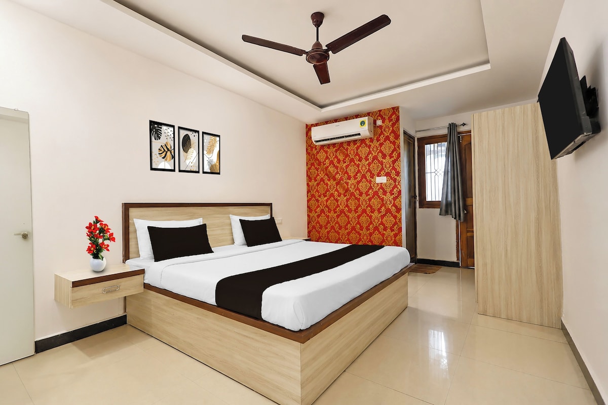 A hotel room with a bed, a nightstand, a vase with flowers, a ceiling fan, an air conditioning unit, and a television mounted on the wall.