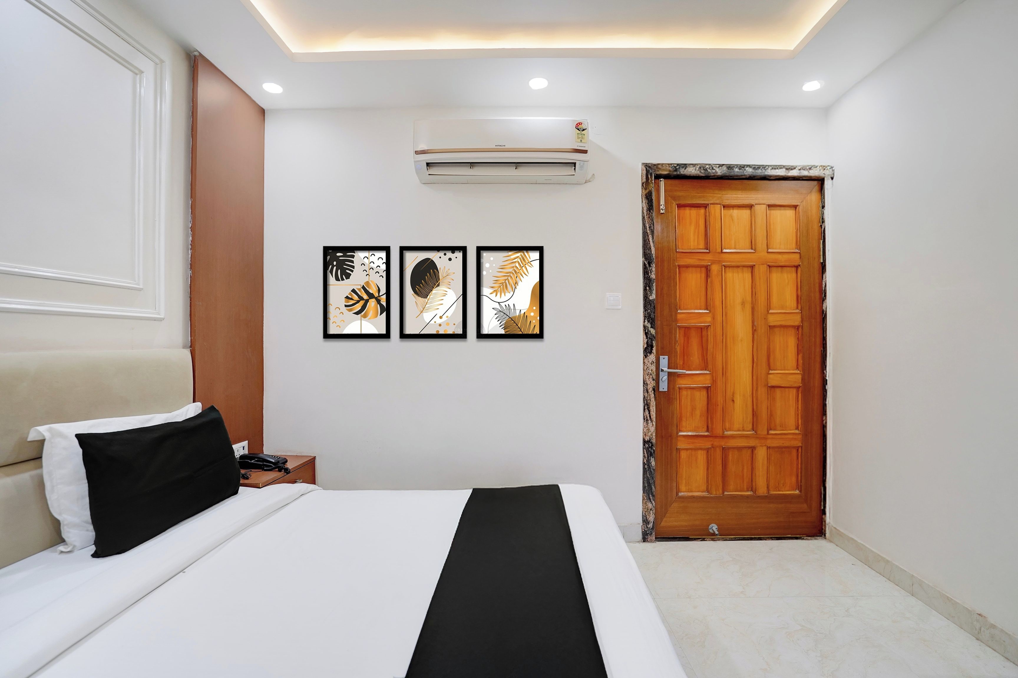 Une chambre d’hôtel avec un lit, une table de chevet avec un téléphone, une porte en bois et trois œuvres d’art encadrées accrochées au mur.