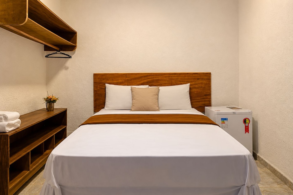 Quarto de hotel com cabeceira de madeira, roupa de cama branca, mesa de cabeceira com abajur e um pequeno frigorífico.