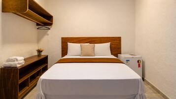 A hotel room with a wooden headboard, white bedding, a bedside table with a lamp, and a small refrigerator.