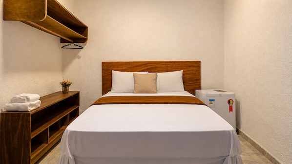 A hotel room with a wooden headboard, white bedding, a bedside table with a lamp, and a small refrigerator.